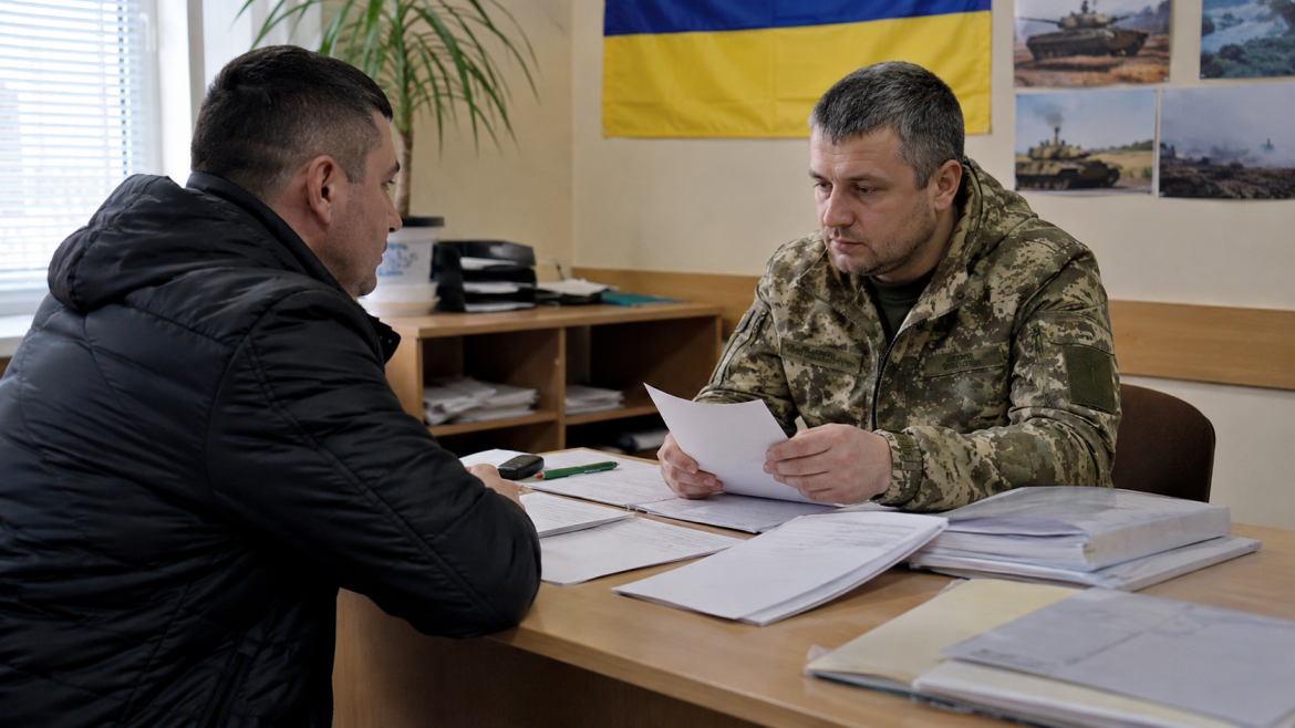 Ultra-realistic documentary photography of a Ukrainian man of Slavic appearance consulting with an official at a military registration office, documents on the desk without readable text, calm administrative atmosphere, concept of mobilization rules, age categories and legal procedures in Ukraine in 2026, realistic people and details, bright vivid colors, natural lighting, professional documentary photography style, high detail, no readable text, no letters, no logos, no watermark, 16:9 aspect ratio
